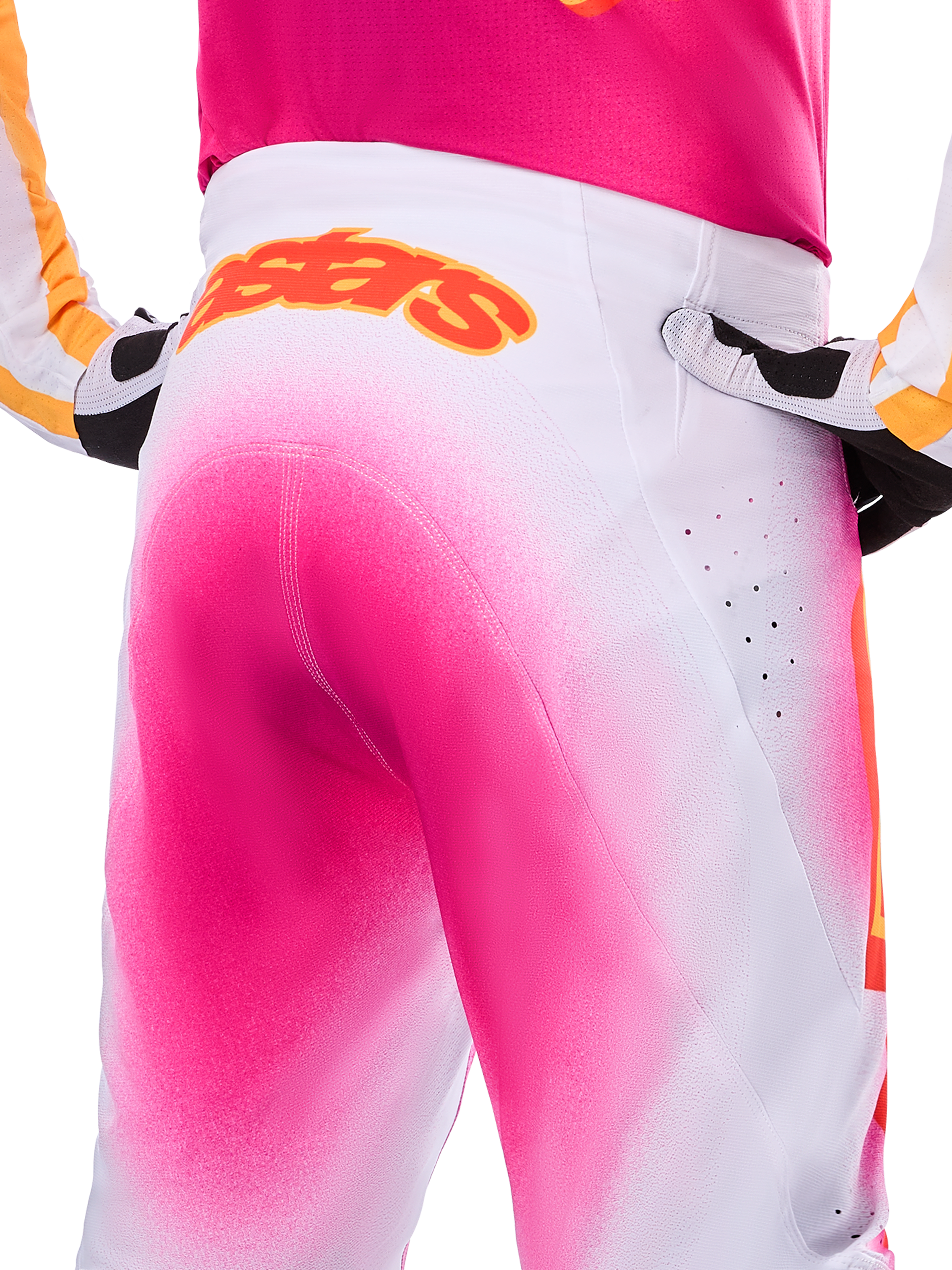 Alpinestars Limited Edition Supertech AFD MIG Pants, pantaloni da motocross, rosa fluo e bianco con un effetto sfumato a spruzzo, grande logo Alpinestars arancione sul girovita posteriore, vista ravvicinata che mostra la vestibilità atletica e le perforazioni per la ventilazione.
