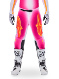 Alpinestars Limited Edition Supertech AFD MIG Pants, pantaloni da motocross, rosa fluo e bianco con accenti arancioni, caratterizzati da grandi loghi a stella sulle cosce, pannelli perforati e zone di grip nere sulle ginocchia, indossati con stivali da motocross bianchi e neri
