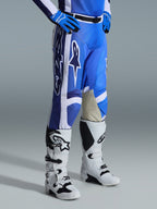 Pantaloni Alpinestars 2026 Racer Air Portl, pantaloni da motocross, blu Ucla e bianco, indossati con guanti blu coordinati e stivali da motocross bianchi e neri, con grandi loghi Alpinestars bianchi, pannelli interni alle ginocchia in pelle beige e fibbia magnetica nera in vita.