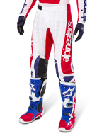 Alpinestars Limited Edition Techstar Fortitude Pants, pantaloni da motocross, rosso acceso, bianco e blu, indossati con stivali e guanti da fuoristrada coordinati, caratterizzati da marchio verticale sulla gamba, pannelli traforati traspiranti e sezioni rinforzate nere sulle ginocchia, design con vestibilità atletica da gara.