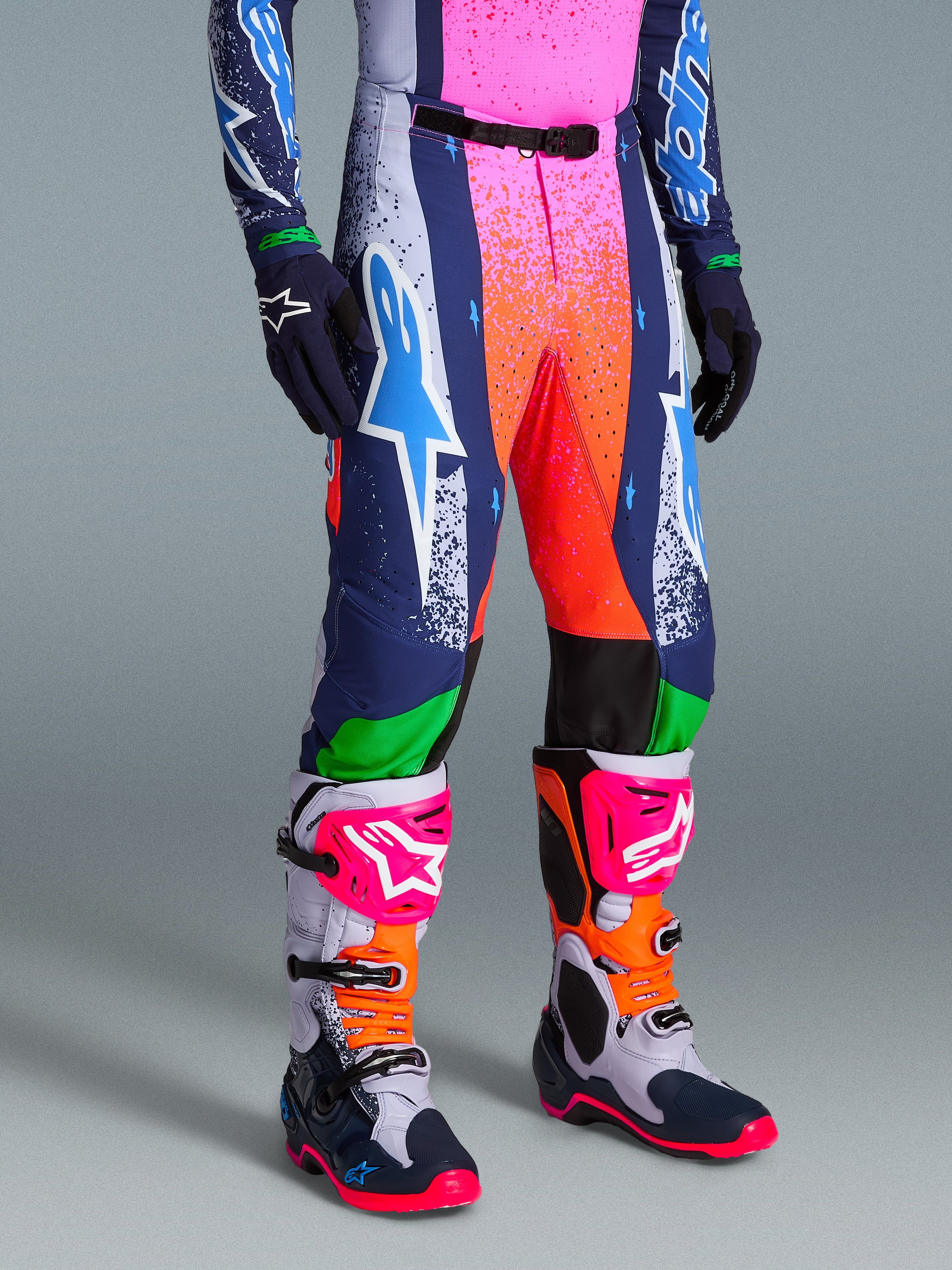 Alpinestars Pantaloni Supertech Records Limited Edition, vivaci pantaloni da motocross multicolore con grafiche a spruzzo rosa e arancioni, pannelli blu navy e bianchi, con un grande logo Astars bianco e ciano, indossati con stivali e guanti tecnici coordinati.