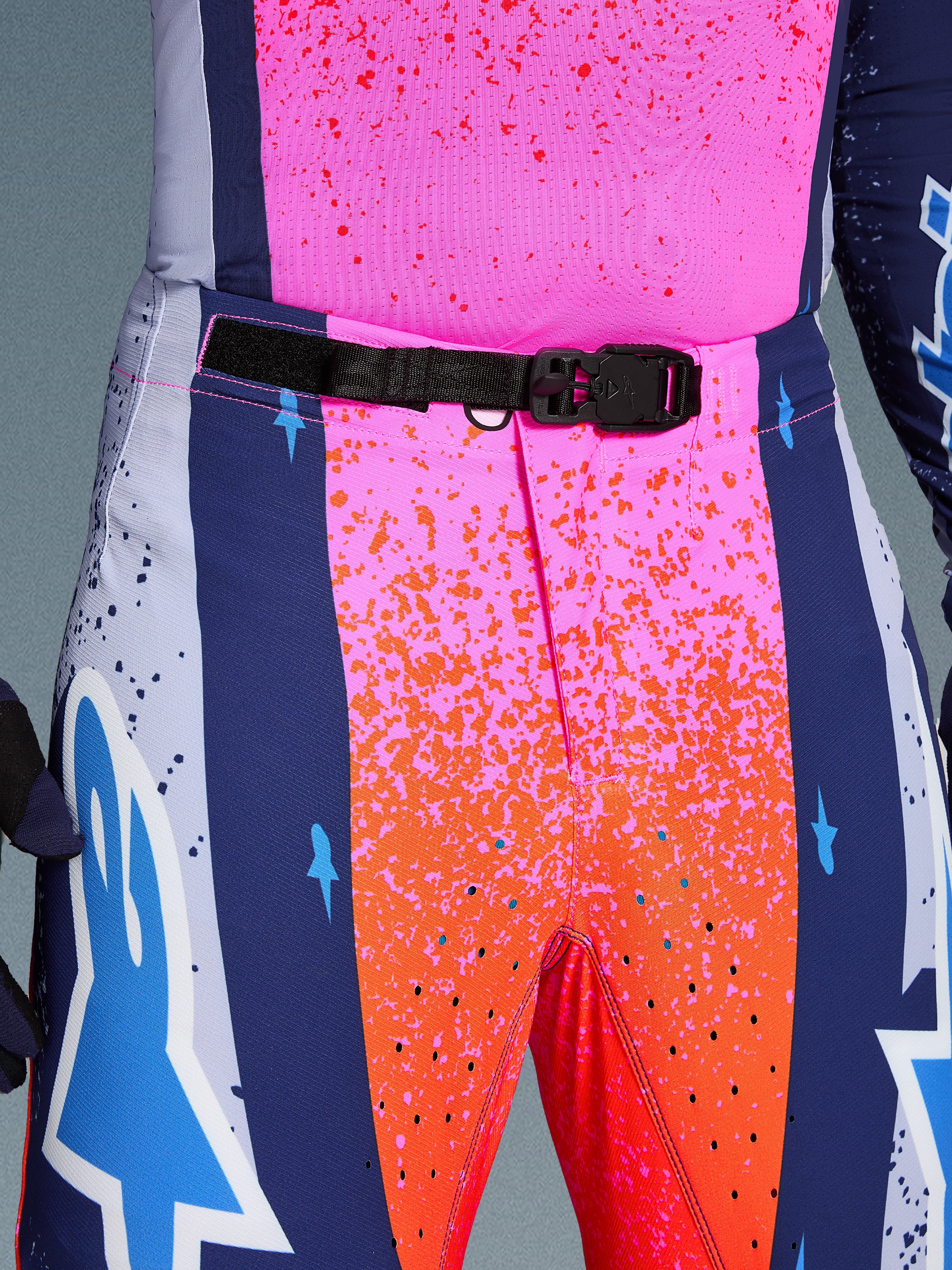 Alpinestars Limited Edition Supertech Records Pants, pantaloni da motocross, multicolore rosa e arancione con pannelli laterali blu navy, primo piano della vita con fibbia magnetica SX1 nera, tessuto rip-stop con grafiche splatter e perforazioni laser.