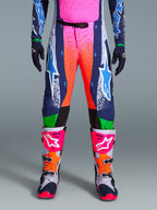 Alpinestars Limited Edition Supertech Records Pants, pantaloni da motocross, multicolore con vivaci grafiche splash rosa e arancione, pannelli blu navy con loghi a stella blu e accenti verdi, indossati con maglia coordinata e stivali da motocross grigi, rosa e arancioni.