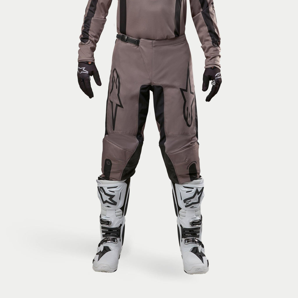 Pantaloni Alpinestars Fluid Lurv 2024, pantaloni da motocross, Mud Black, indossati con maglia coordinata, guanti neri e stivali bianchi, caratterizzati da grandi loghi neri sulle cosce, vestibilità ergonomica e materiale resistente per corse fuoristrada