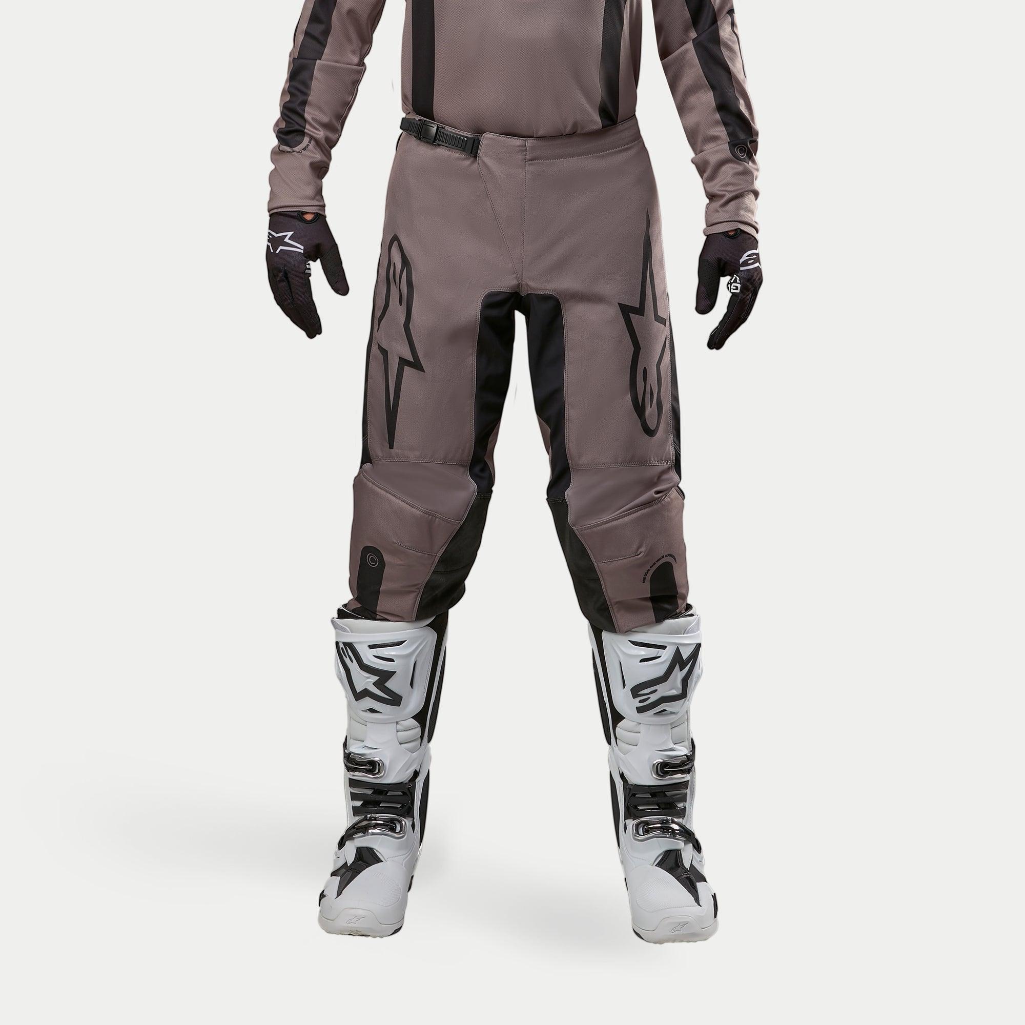 Pantaloni Alpinestars Fluid Lurv 2024, pantaloni da motocross, Mud Black, indossati con maglia coordinata, guanti neri e stivali bianchi, caratterizzati da grandi loghi neri sulle cosce, vestibilità ergonomica e materiale resistente per corse fuoristrada