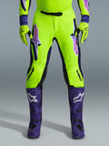 Alpinestars Pantaloni Supertech Vista 2026, pantaloni da motocross, giallo fluorescente, viola e nero, indossati con guanti coordinati e stivali viola, con grandi loghi sfumati sulle cosce, zone perforate per la ventilazione e design atletico professionale per gare off-road