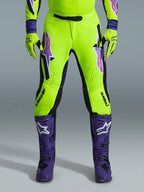 Alpinestars Pantaloni Supertech Vista 2026, pantaloni da motocross, giallo fluorescente, viola e nero, indossati con guanti coordinati e stivali viola, con grandi loghi sfumati sulle cosce, zone perforate per la ventilazione e design atletico professionale per gare off-road