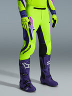 Alpinestars pantaloni Supertech Vista 2026, pantaloni da motocross, giallo neon brillante, viola e nero, indossati con stivali viola coordinati e guanti gialli, caratterizzati da un logo evidente sulla coscia e una vestibilità atletica performante.