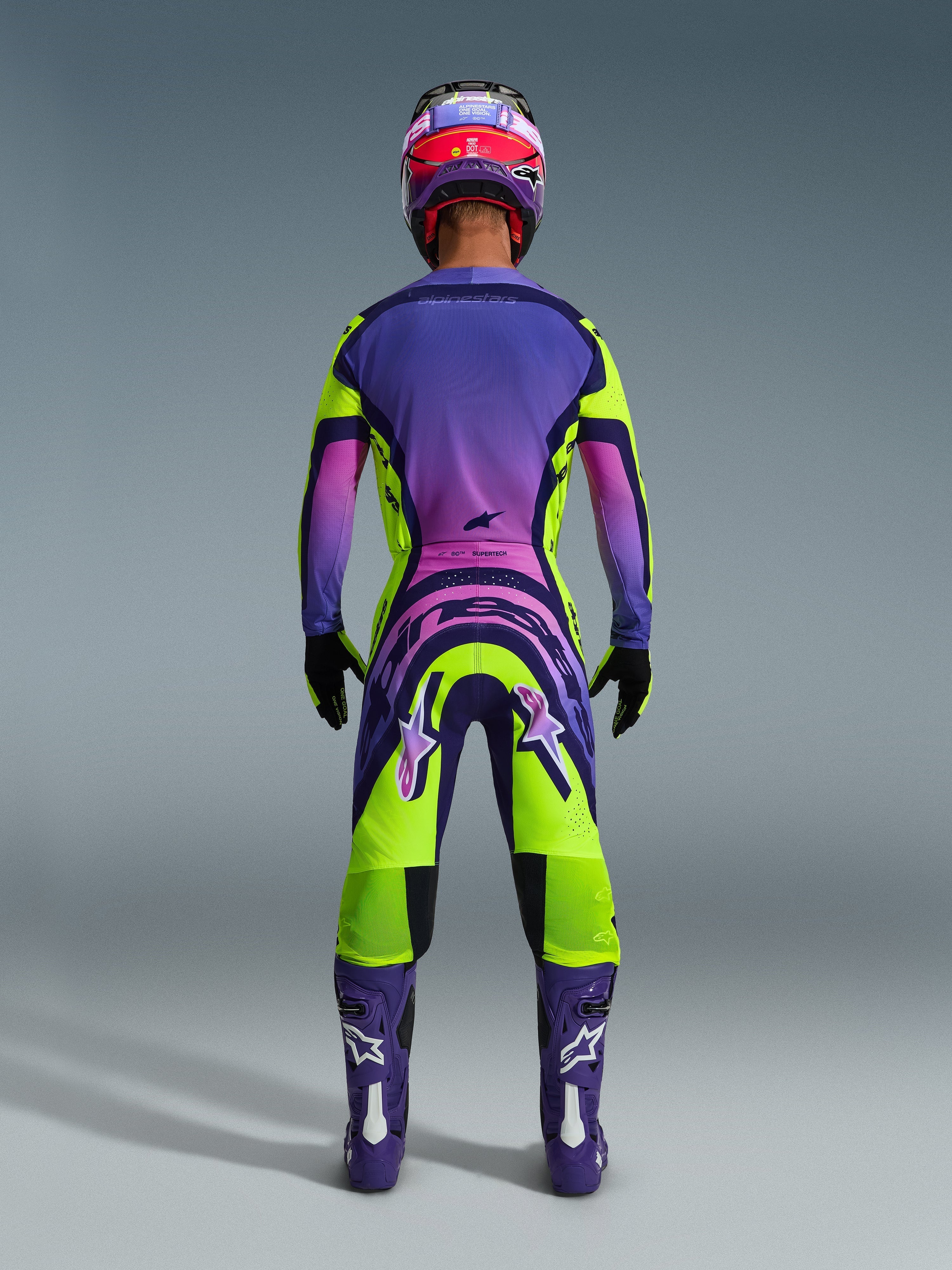 Pilota di motocross visto da dietro che indossa la maglia Supertech Vista 2026 e i pantaloni coordinati, design giallo e viola vibrante con sfumature rosa, stivali viola con loghi Alpinestars bianchi, guanti neri e casco da corsa rosso e viola, set di abbigliamento professionale da gara.