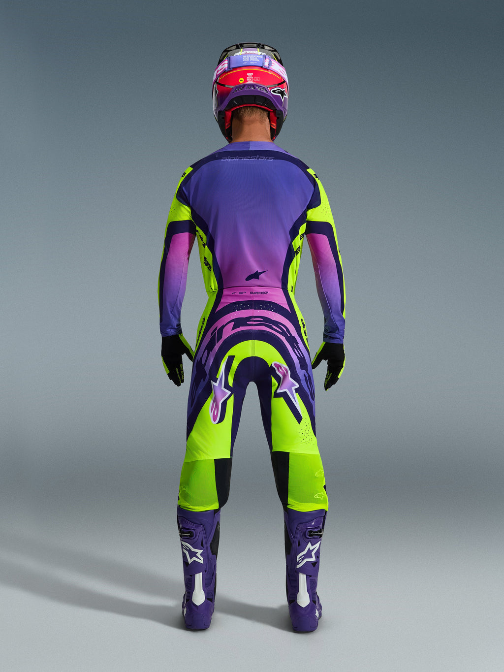 Set abbigliamento Alpinestars 2026 Supertech Vista, vista posteriore di un pilota di motocross con completo da gara sfumato in viola vivace, rosa e verde lime, maglia e pantaloni coordinati con loghi evidenti, casco coordinato viola e rosa, stivali viola e guanti neri.