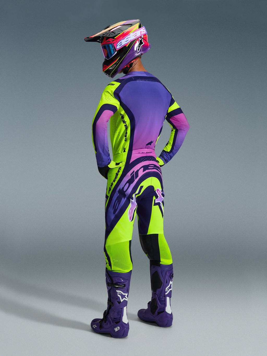 Pilota di motocross visto di spalle che indossa il completo Alpinestars Supertech Vista 2026, maglia viola con sfumatura rosa e accenti giallo neon, pantaloni coordinati giallo neon e viola, stivali viola scuro con loghi bianchi, casco e maschera coordinati, abbigliamento da motocross di livello professionale