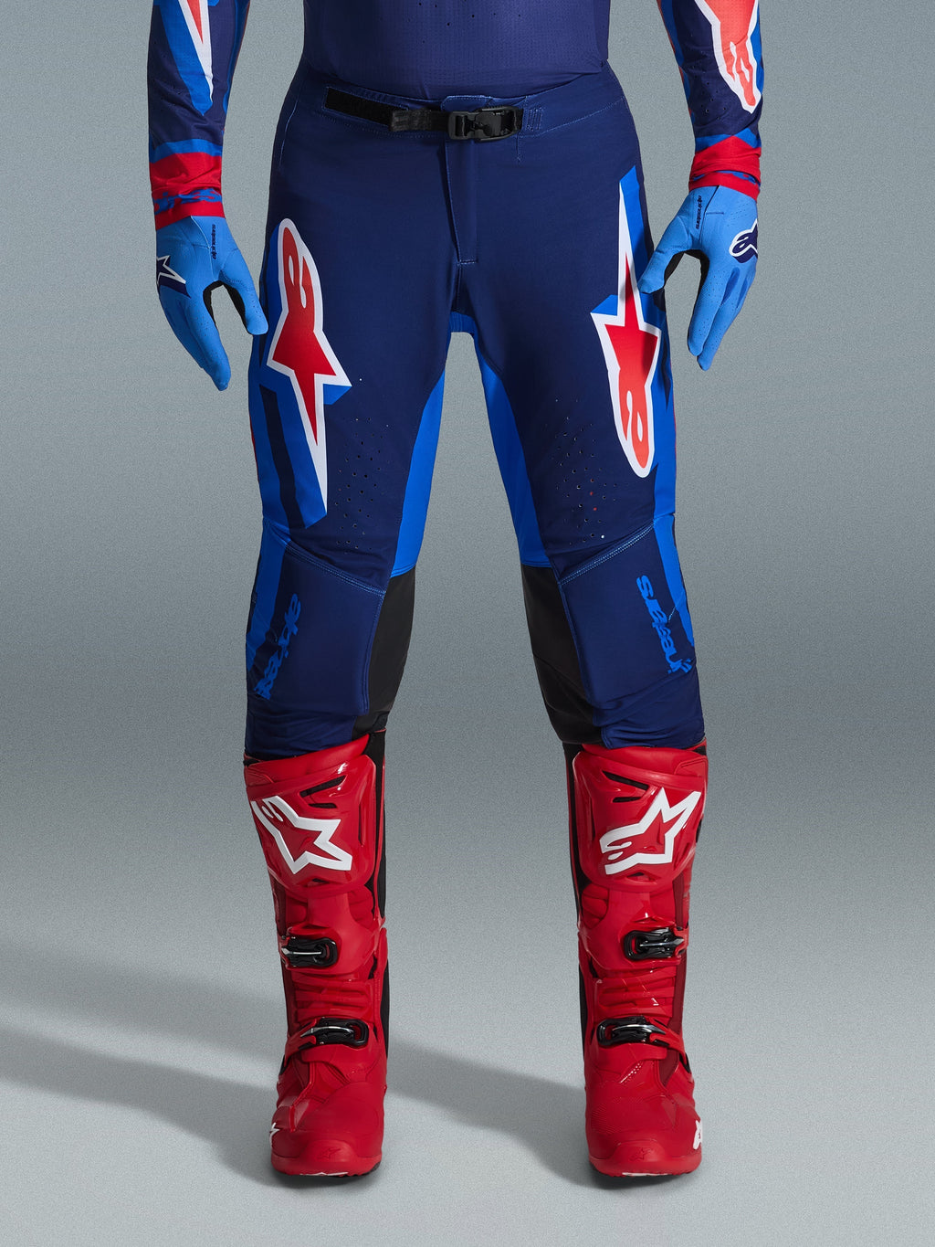 Alpinestars Pantaloni Supertech Vista 2026, pantaloni da motocross, blu scuro, corallo acceso e cobalto, vista frontale del pilota che indossa abbigliamento tecnico con guanti blu coordinati e stivali da motocross rossi, caratterizzati da vistosi loghi Alpinestars rossi e bianchi sulle cosce.