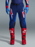 Alpinestars Pantaloni Supertech Vista 2026, pantaloni da motocross, blu scuro, corallo acceso e cobalto, vista frontale del pilota che indossa abbigliamento tecnico con guanti blu coordinati e stivali da motocross rossi, caratterizzati da vistosi loghi Alpinestars rossi e bianchi sulle cosce.