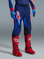 Alpinestars Supertech Vista Pants 2026, pantaloni da motocross, Dark Navy Hot Coral Cobalt, indossati con stivali da gara rosso acceso e guanti blu, con grandi loghi grafici sulle cosce e pannelli interni rinforzati neri per prestazioni fuoristrada