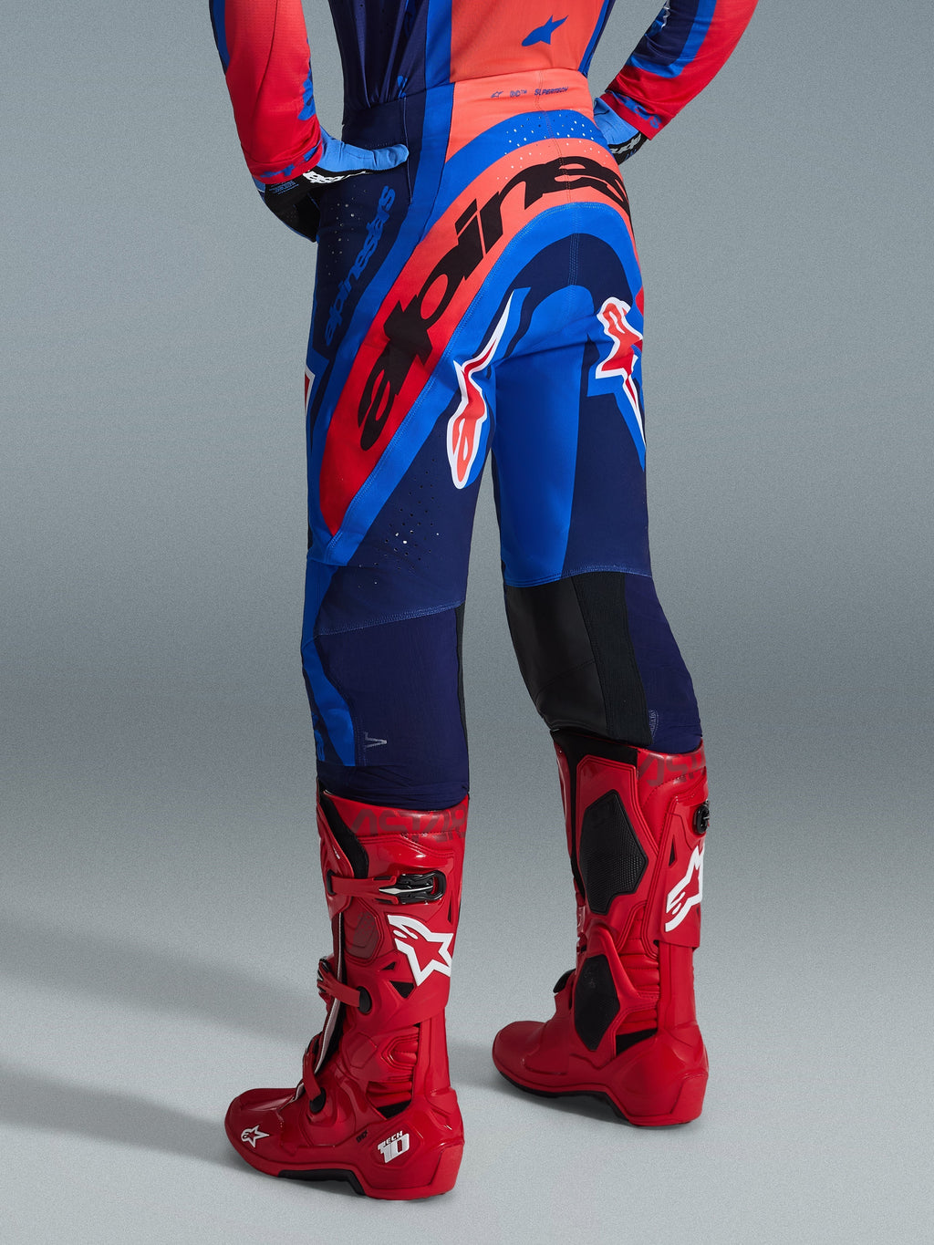 Alpinestars 2026 Supertech Vista Pants, pantaloni da motocross, Dark Navy Hot Coral Cobalt, vista posteriore con vistoso marchio Alpinestars rosso sulla seduta, abbinati a stivali da motocross rosso brillante, costruzione leggera ed elasticizzata per competizioni professionistiche