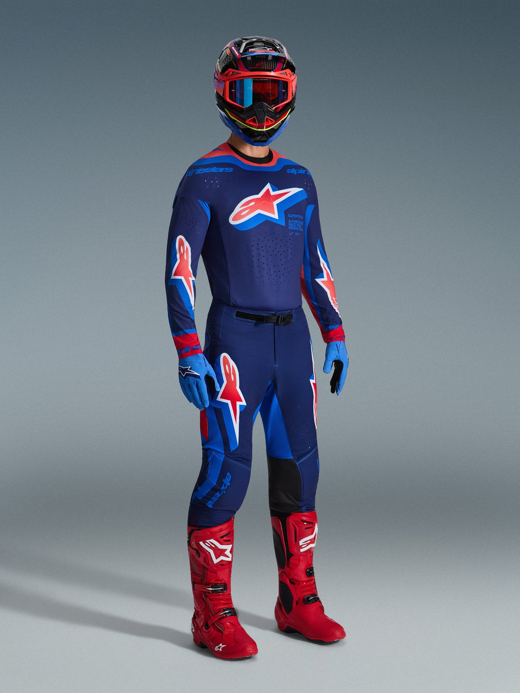 Pilota di motocross che indossa pantaloni Supertech Vista 2026 in Dark Navy Hot Coral Cobalt, maglia da gara coordinata a maniche lunghe, stivali da motocross rossi e casco coordinato con occhiali rossi, scatto in studio a figura intera di abbigliamento tecnico da fuoristrada