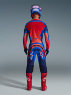 Alpinestars 2026 Supertech Vista Pants, pantaloni da motocross, Dark Navy Hot Coral e Cobalt, vista posteriore di un pilota in equipaggiamento da gara completo con maglia coordinata, casco blu e stivali rossi, con branding evidente sulla seduta e sulle gambe.