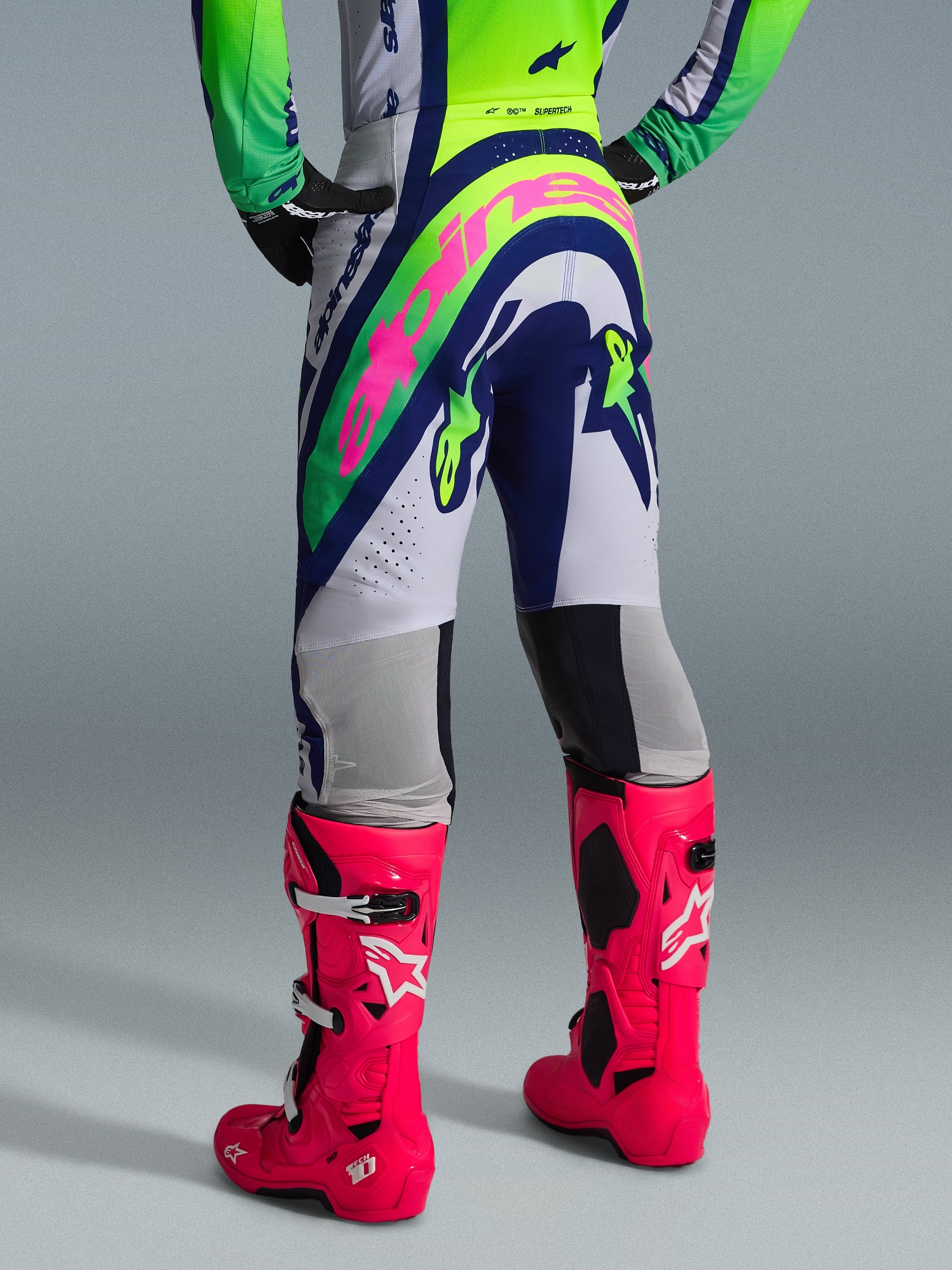 Alpinestars 2026 Supertech Vista Pants, pantaloni da motocross, grigio chiaro, giallo fluo e blu, vista posteriore con grande logo Alpinestars rosa, indossati con maglia da gara coordinata e stivali Tech 10 rosa acceso, equipaggiamento professionale da fuoristrada