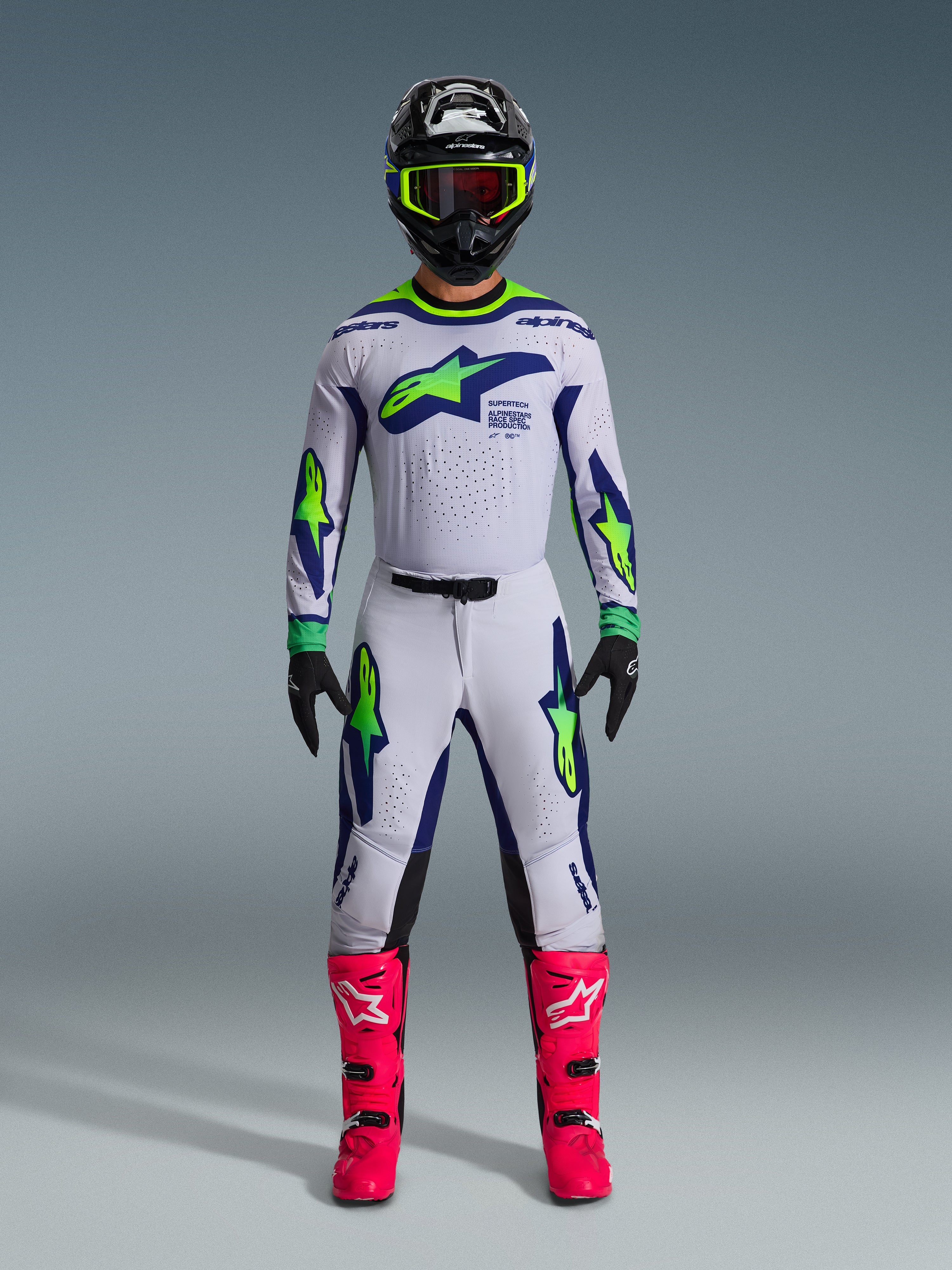 Alpinestars Set Completo Supertech Vista 2026, pilota di motocross con abbigliamento da gara completo, maglia e pantaloni bianchi con dettagli blu e verde neon, casco nero con finiture giallo neon, stivali rosa acceso, completo professionale per gare off-road