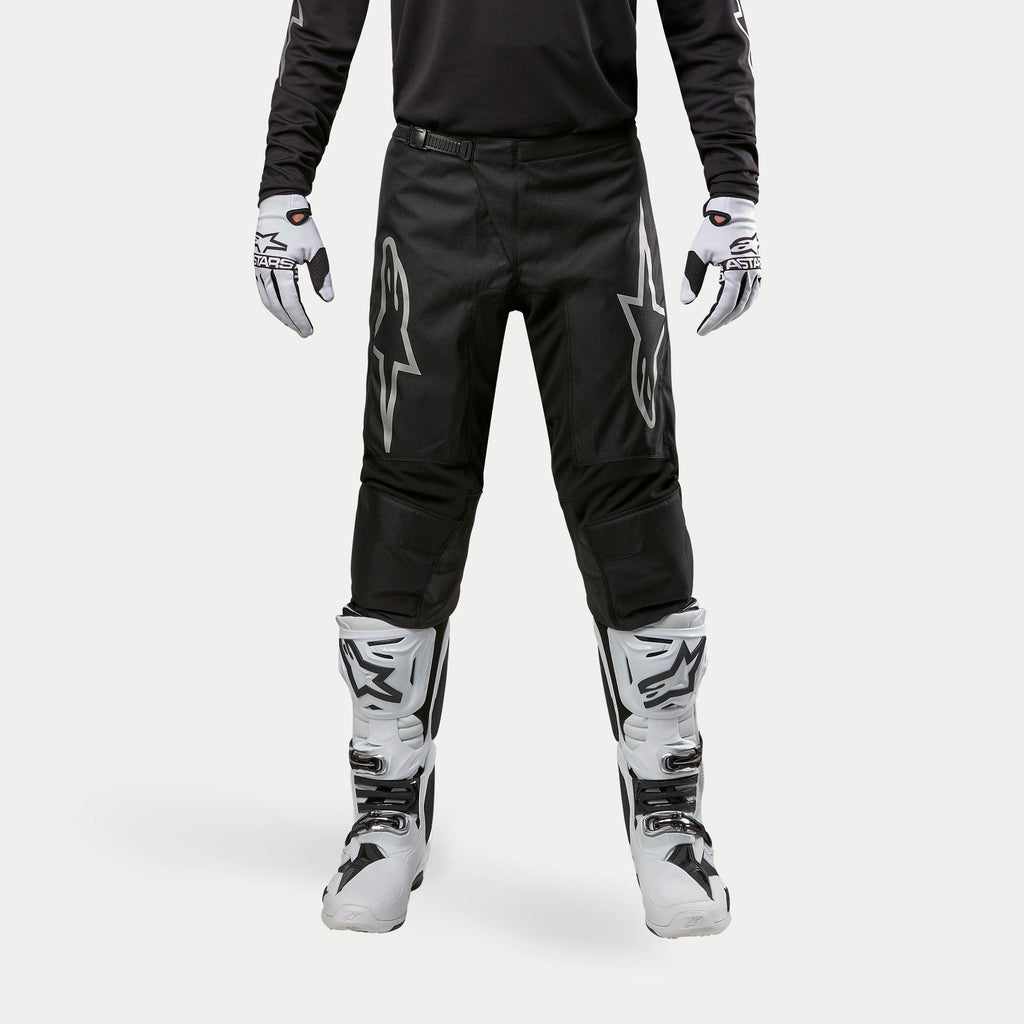 Alpinestars 2024 Fluid Graphite Pants, pantaloni da motocross, nero e argento, con loghi Alpinestars argento sulle cosce, indossati con stivali e guanti da motocross bianchi e neri, design sportivo per corse fuoristrada