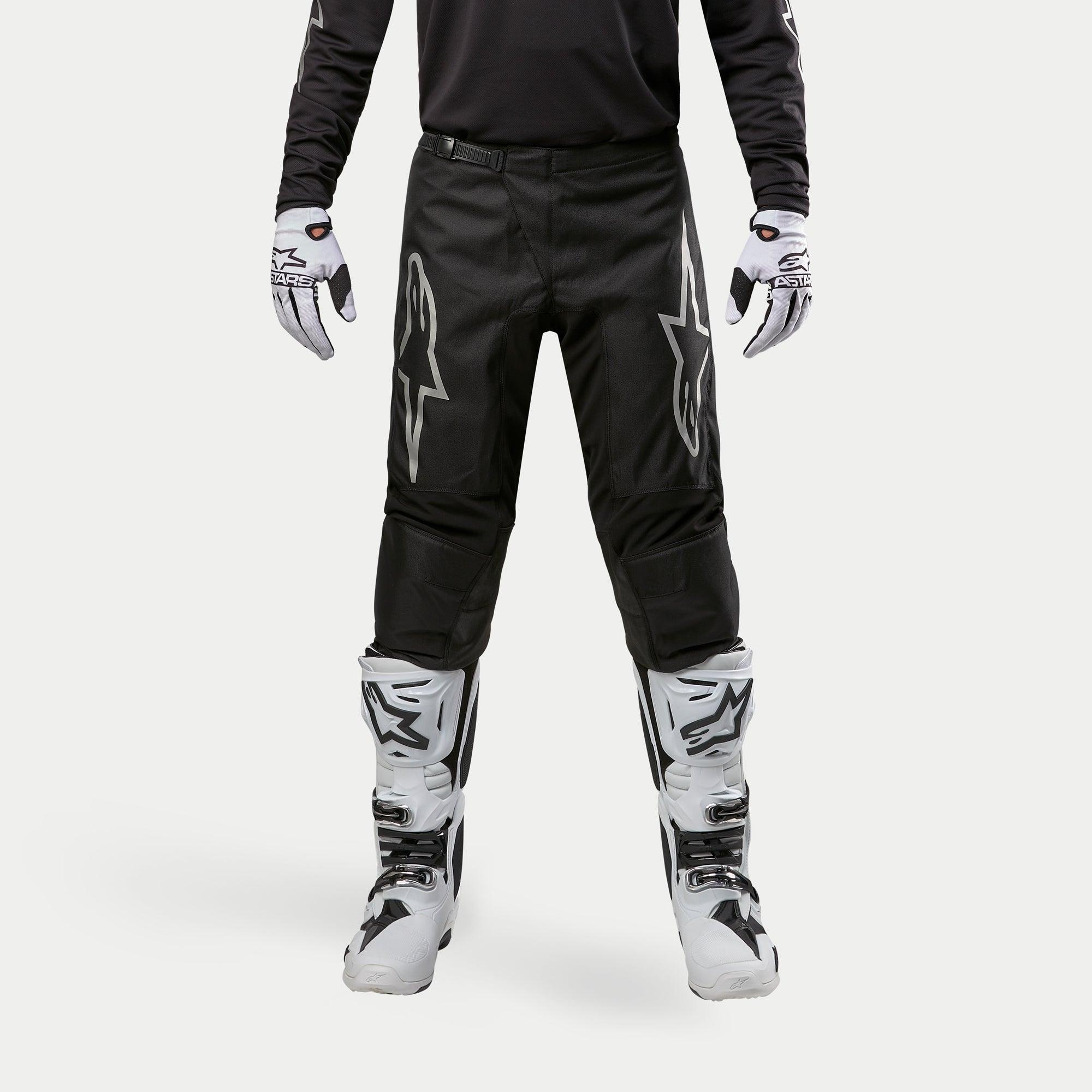 Alpinestars 2024 Fluid Graphite Pants, pantaloni da motocross, nero e argento, con loghi Alpinestars argento sulle cosce, indossati con stivali e guanti da motocross bianchi e neri, design sportivo per corse fuoristrada