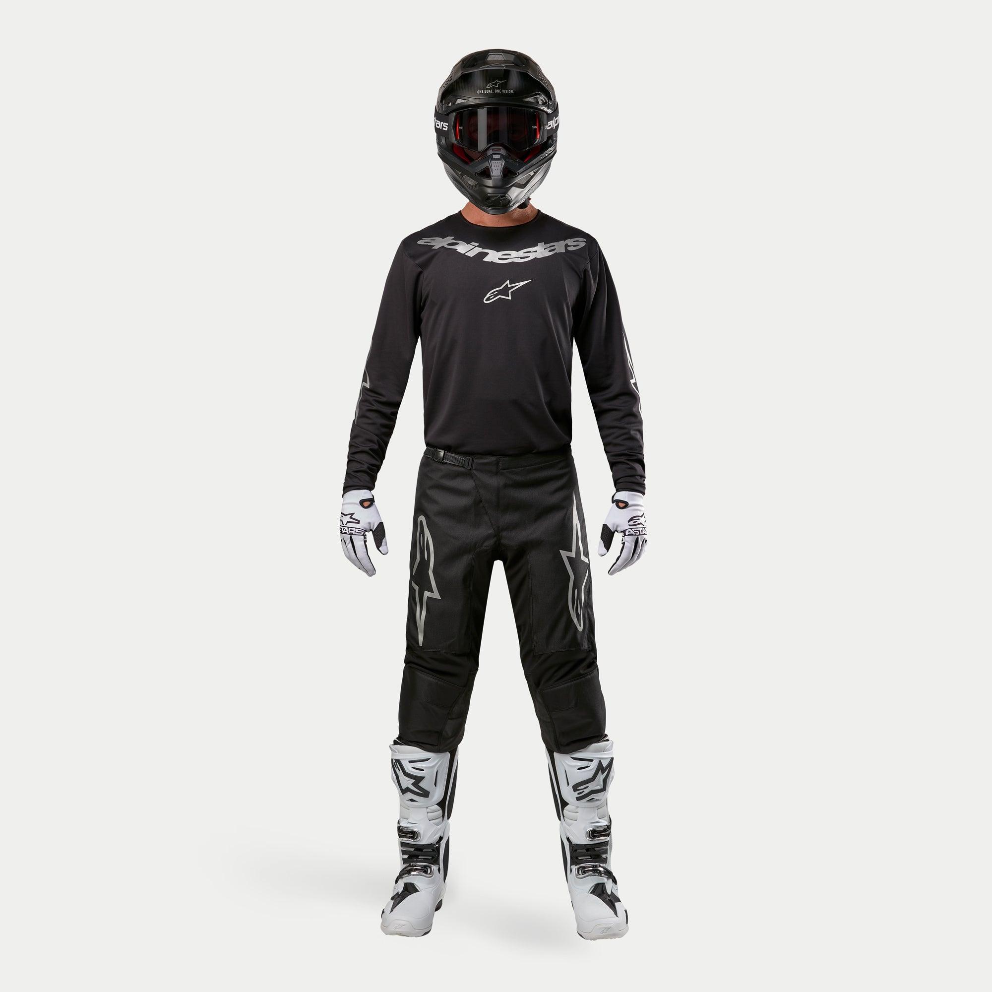 Pilota di motocross con equipaggiamento da gara completo, pantaloni Alpinestars Fluid Graphite 2024 in nero e argento, maglia nera a maniche lunghe con loghi argento, casco nero con occhiali, stivali bianchi e neri, guanti bianchi e neri, completo coordinato da motocross