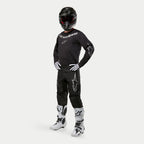 Alpinestars 2024 Fluid Graphite Pants, pantaloni da motocross, nero e argento, indossati con maglia nera coordinata, casco nero, guanti bianchi e stivali bianchi, con loghi Alpinestars argento sulle cosce, vestibilità atletica ergonomica per corse fuoristrada.
