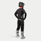 Alpinestars Pantaloni Fluid Graphite 2024, pantaloni da motocross, nero e argento, vista posteriore su un pilota che indossa una maglia nera, un casco nero e stivali bianchi e neri, con branding argento sulla parte bassa della schiena e una struttura in poli-tessuto resistente per le gare fuoristrada
