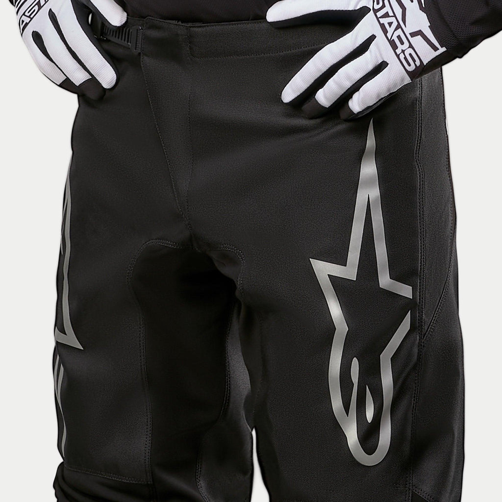 Alpinestars Pantaloni Fluid Graphite 2024, pantaloni da motocross, nero e argento, primo piano della parte superiore delle cosce e della vita, con un grande logo a stella argento sulla gamba, indossati con guanti da gara bianchi e neri