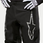 Alpinestars Pantaloni Fluid Graphite 2024, pantaloni da motocross, nero e argento, primo piano della parte superiore delle cosce e della vita, con un grande logo a stella argento sulla gamba, indossati con guanti da gara bianchi e neri