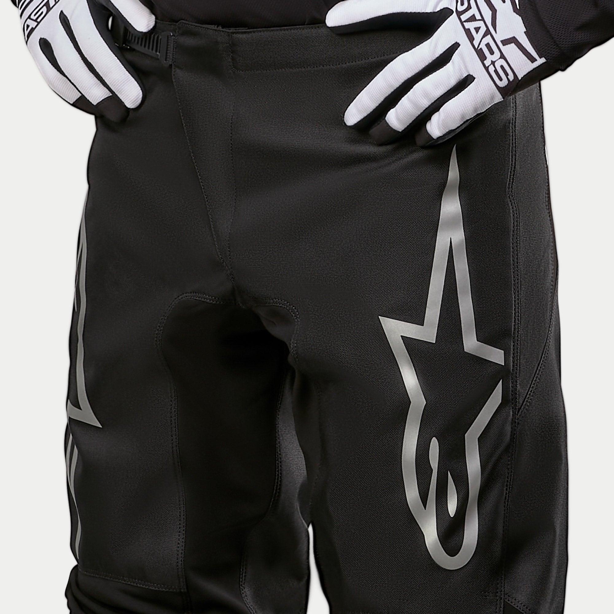 Alpinestars Pantaloni Fluid Graphite 2024, pantaloni da motocross, nero e argento, primo piano della parte superiore delle cosce e della vita, con un grande logo a stella argento sulla gamba, indossati con guanti da gara bianchi e neri