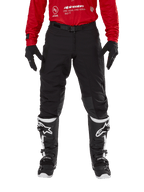 Pantaloni Alpinestars Techdura, pantaloni da enduro e off-road, nero, indossati con una maglia Alpinestars rossa e stivali bianchi e neri, caratterizzati da una robusta costruzione in tela elasticizzata, fibbia magnetica regolabile e rinforzi in pelle sulle ginocchia per prestazioni fuoristrada.
