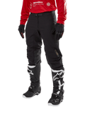 Alpinestars Techdura Pants, pantaloni da moto fuoristrada neri, indossati con una maglia rossa a maniche lunghe e stivali da motocross bianchi e neri, caratterizzati da una robusta costruzione in poliammide elasticizzata, rinforzi interni in pelle sulle ginocchia e cintura regolabile con fibbia per la guida enduro.