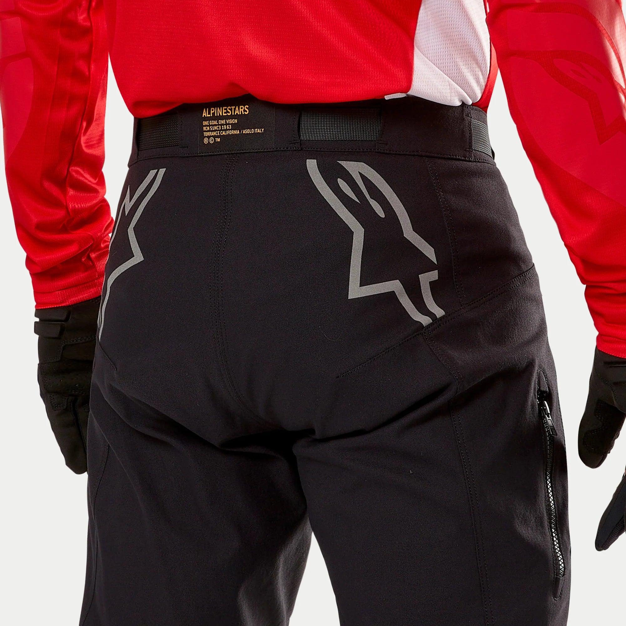 Alpinestars Techdura Pants, pantaloni da enduro neri, vista posteriore con grandi loghi Astars grigi sulla seduta, indossati con maglia rossa e guanti neri, materiale resistente con zip laterale e vita logata