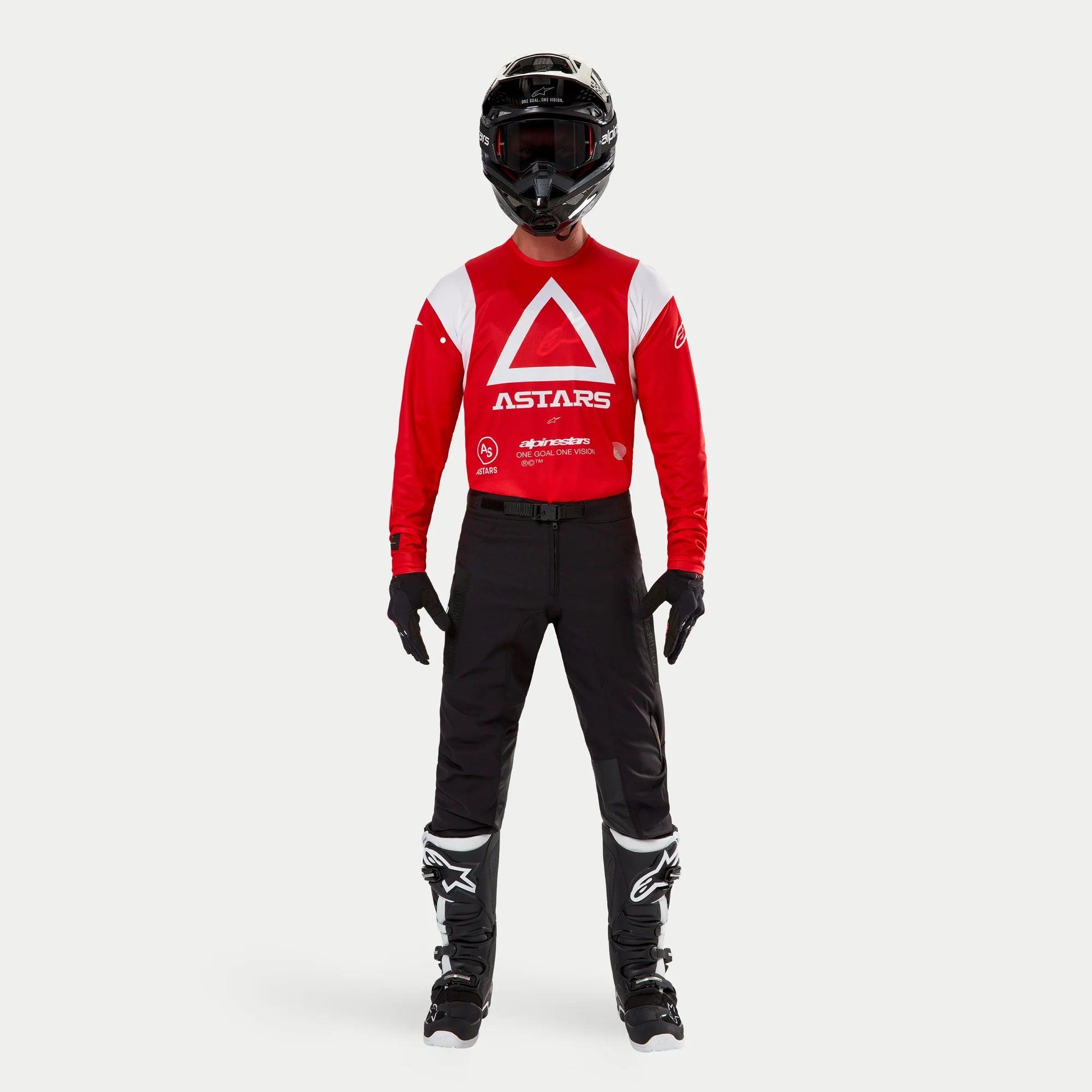 Alpinestars Techdura Pants, pantaloni da enduro e off-road, nero, indossati con maglia a maniche lunghe Astars rossa e bianca, casco nero e stivali neri e bianchi, caratterizzati da una robusta costruzione in poliammide elasticizzata e pannelli tecnici per la guida off-road.