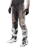 Alpinestars 2024 Techstar Pneuma Pants, pantaloni da motocross, sabbia scura e grigio polvere di ferro, indossati con stivali bianchi e neri e guanti neri, caratterizzati da un motivo ondulato con un grande logo a stella, abbigliamento resistente per corse fuoristrada