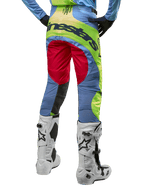 Alpinestars 2024 Techstar Ocuri Pants, pantaloni da motocross, celeste, giallo fluo e rosso berry, vista posteriore con stivali da motocross bianchi e neri, caratterizzati da vivaci blocchi di colore e vistoso marchio Alpinestars sulla seduta.
