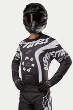 Alpinestars 2024 Racer Hana Jersey, maglia da motocross, nero e bianco, con grafiche testuali e logo Alpinestars in bianco, indossata da un pilota con casco nero e maschera, abbigliamento ad alte prestazioni per corse fuoristrada