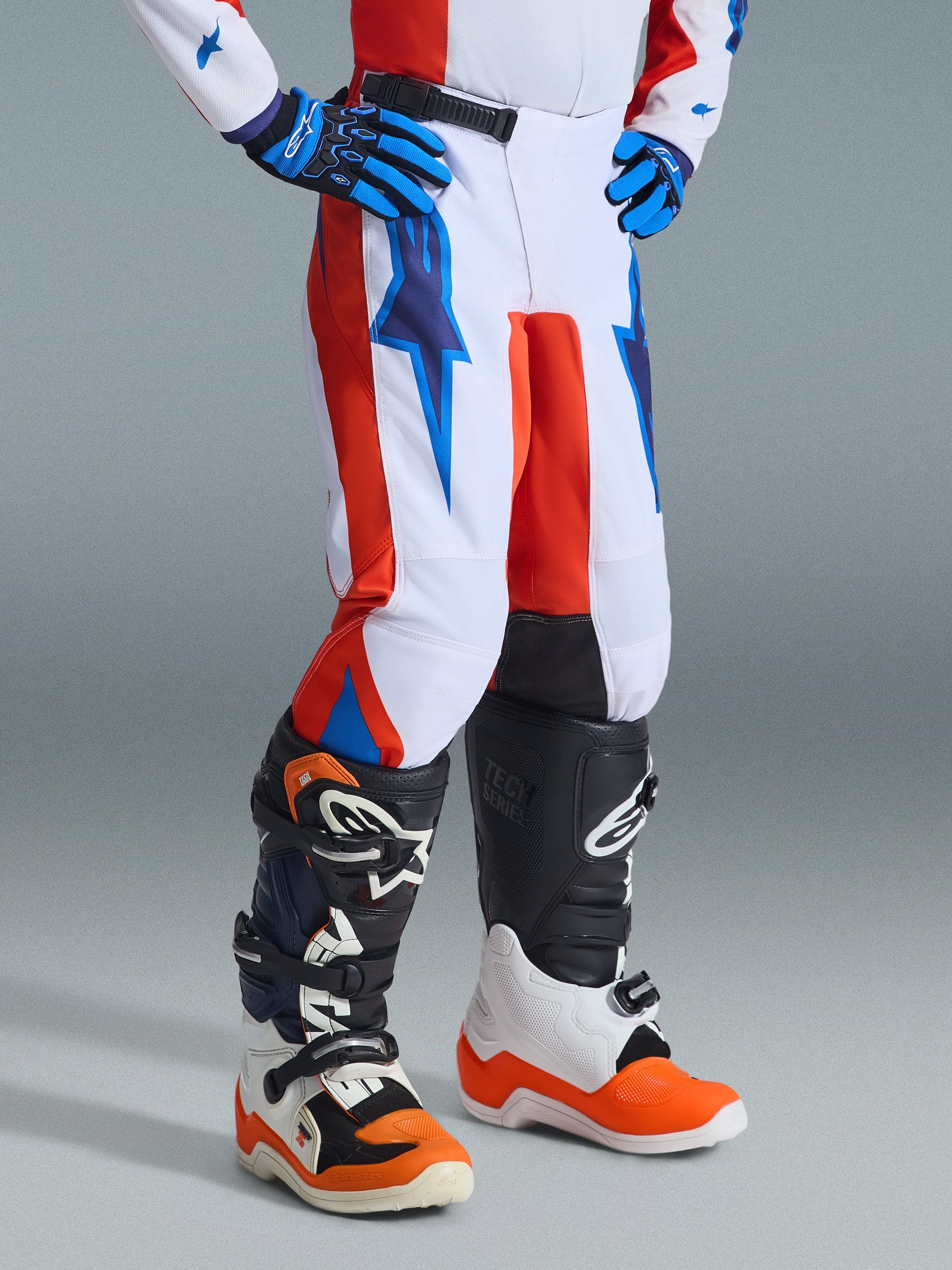 Alpinestars pantaloni Youth Fluid Grid 2026, pantaloni da motocross, arancione e blu UCLA su bianco, indossati con guanti coordinati e stivali da fuoristrada, con grafiche stile fulmine e struttura resistente per giovani piloti