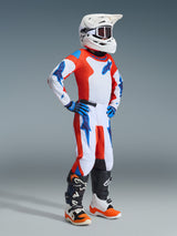 Giovane pilota di motocross con abbigliamento completo da gara, pantaloni e maglia Alpinestars 2026 Youth Fluid Grid in arancione e blu UCLA, casco bianco con occhiali, guanti blu e stivali bianchi e arancioni, outfit coordinato per corse fuoristrada