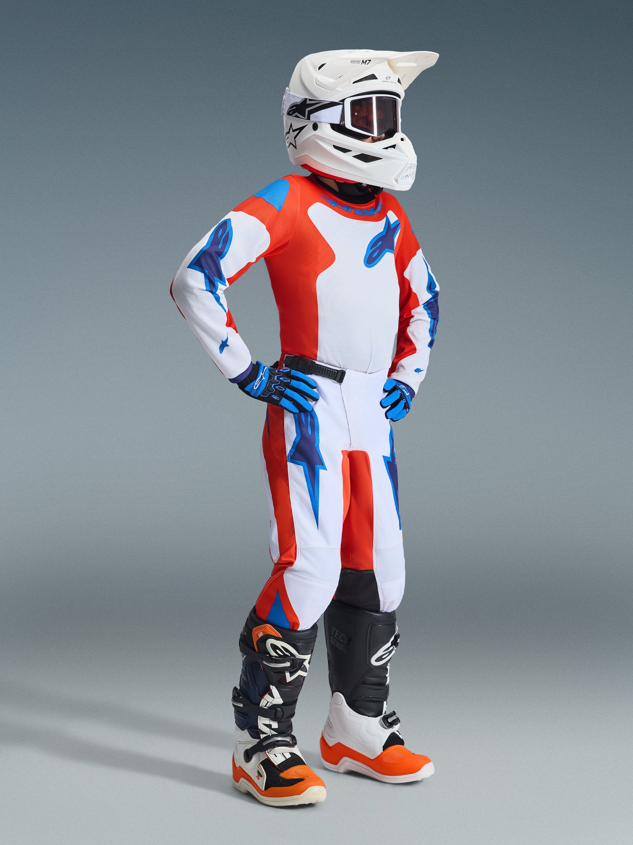 Giovane pilota di motocross con abbigliamento completo da gara, pantaloni e maglia Alpinestars 2026 Youth Fluid Grid in arancione e blu UCLA, casco bianco con occhiali, guanti blu e stivali bianchi e arancioni, outfit coordinato per corse fuoristrada