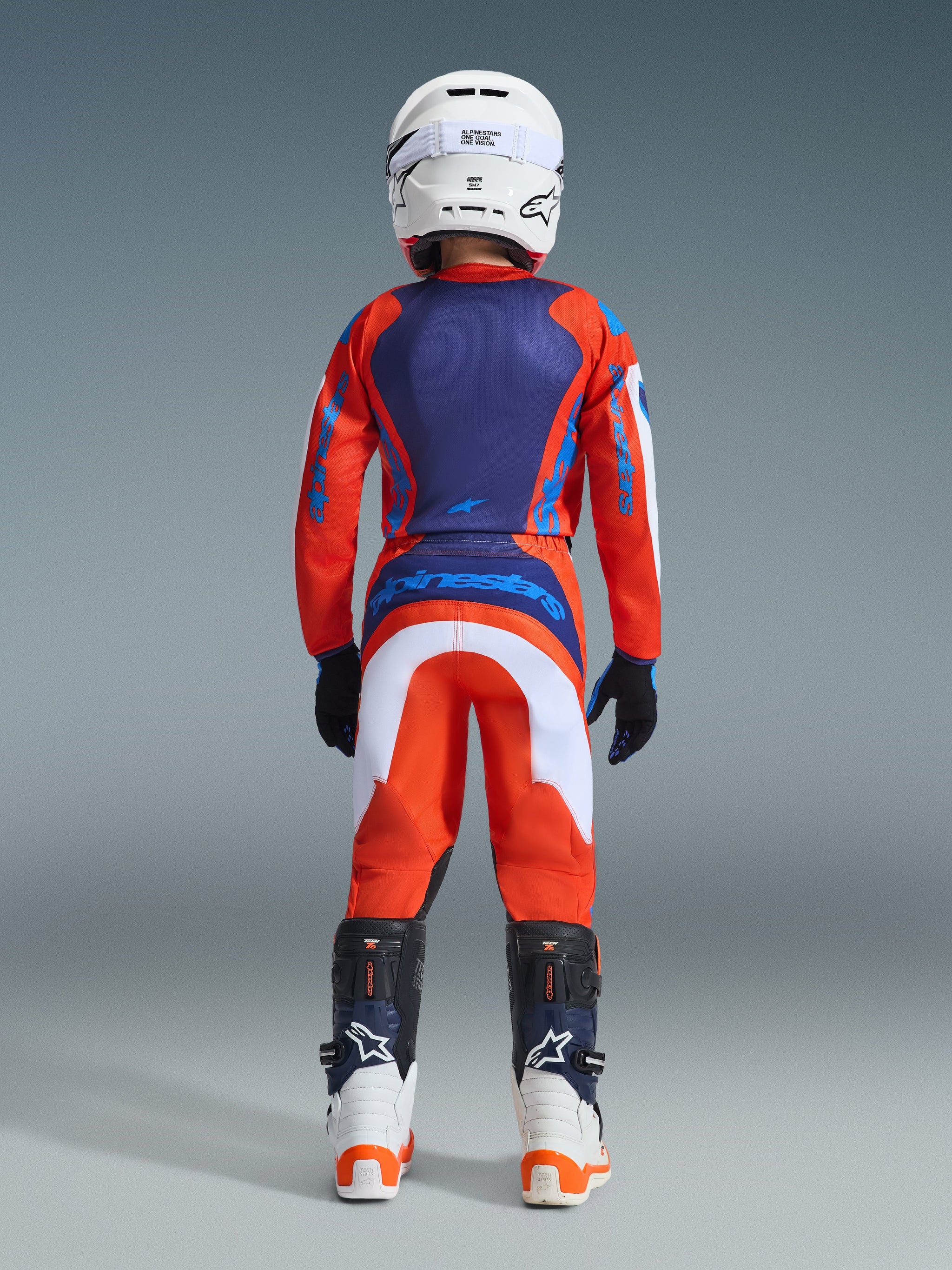 Alpinestars Youth Fluid Grid Pants 2026, pantaloni da motocross, arancione e blu UCLA con accenti bianchi, vista posteriore di un giovane pilota in equipaggiamento completo da gara con maglia coordinata, casco bianco e stivali da motocross bianchi e blu
