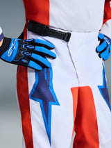 Alpinestars Pantaloni Fluid Grid Ragazzo 2026, pantaloni da motocross, bianco con pannelli arancioni e grafiche a stella blu UCLA, chiusura a cricchetto nera, indossati con guanti coordinati blu e neri, abbigliamento tecnico per ragazzi