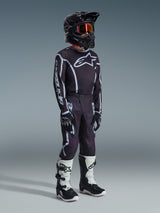 Alpinestars Pantaloni Youth Fluid Apex 2026, pantaloni da motocross, nero e grigio, abbinati a maglia coordinata bianca e nera, casco nero e stivali bianchi e neri, con logo Alpinestars e linee grafiche, completo da gara per motocross giovanile