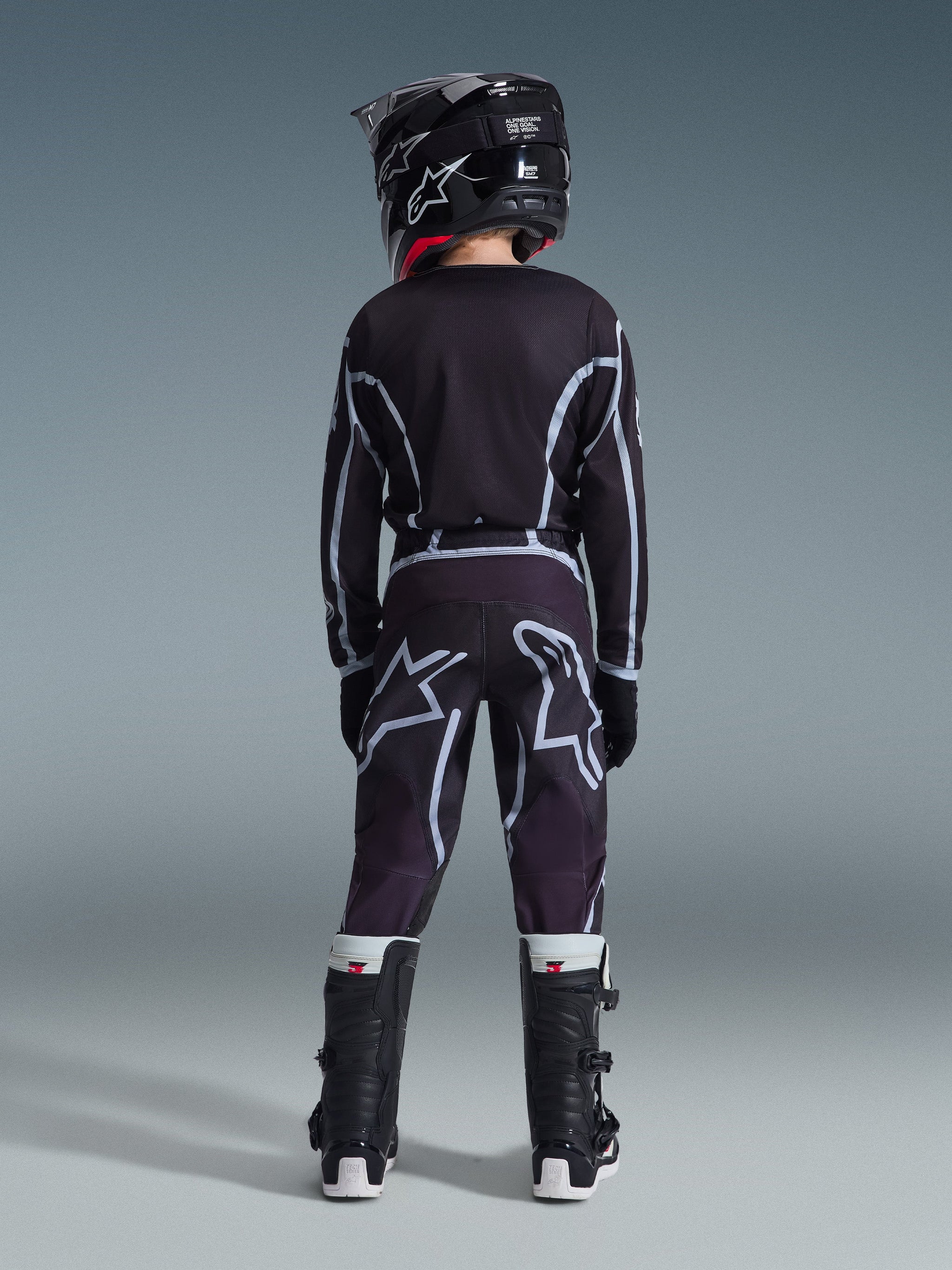 Vista posteriore di un giovane pilota di motocross che indossa i pantaloni Alpinestars 2026 Youth Fluid Apex in nero e grigio, maglia a maniche lunghe coordinata, casco nero e stivali da motocross bianchi e neri, abbigliamento da gara off-road coordinato.