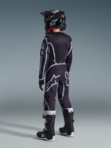 Alpinestars 2026 Pantaloni Youth Fluid Apex, pantaloni da motocross per ragazzi, nero e grigio, indossati da un giovane pilota visti da dietro con maglia e casco coordinati, con loghi bianchi evidenti e motivi geometrici, abbinati a stivali bianchi e neri per corse fuoristrada