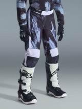 Alpinestars Pantaloni Youth Fluid Lahnd 2026, pantaloni da motocross, mimetica Iron Camo in grigio, nero e bianco, indossati con maglia coordinata, guanti neri e stivali bianchi e neri, con grandi loghi a stella sulle cosce.