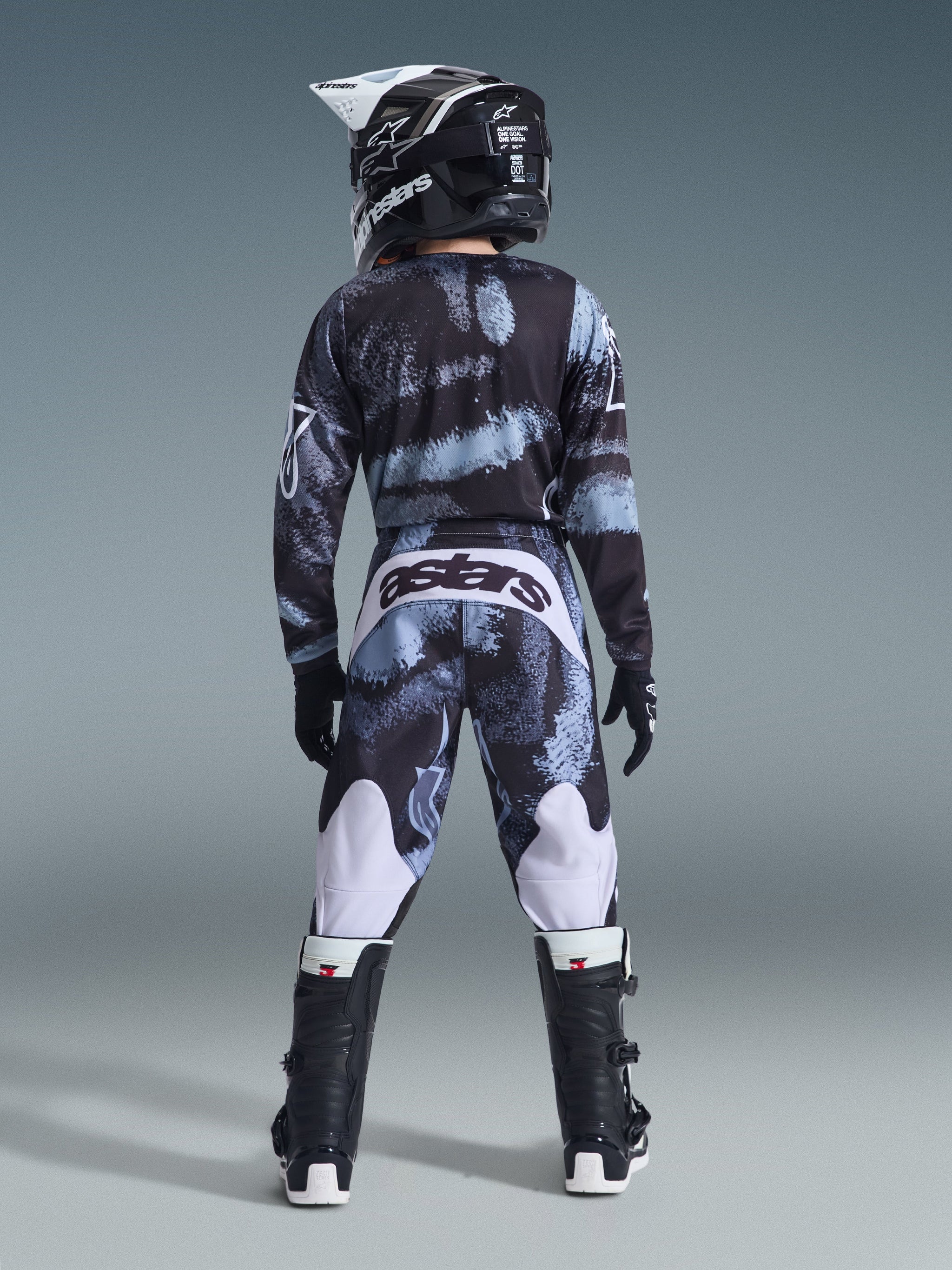 Alpinestars 2026 Youth Fluid Lahnd Pants, pantaloni da motocross, Iron Camo con tonalità blu e grigie, vista posteriore del pilota che indossa il set di abbigliamento coordinato inclusi casco e stivali, accenti bianchi sulle gambe e logo 'astars' sul retro.