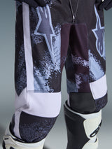 Alpinestars 2026 Youth Fluid Lahnd Pants, pantaloni da motocross, Iron Camo, primo piano della zona delle cosce e delle ginocchia con loghi a stella bianchi, motivo scuro effetto invecchiato con accenti azzurri, indossati con stivali da motocross bianchi e neri, abbigliamento tecnico da fuoristrada