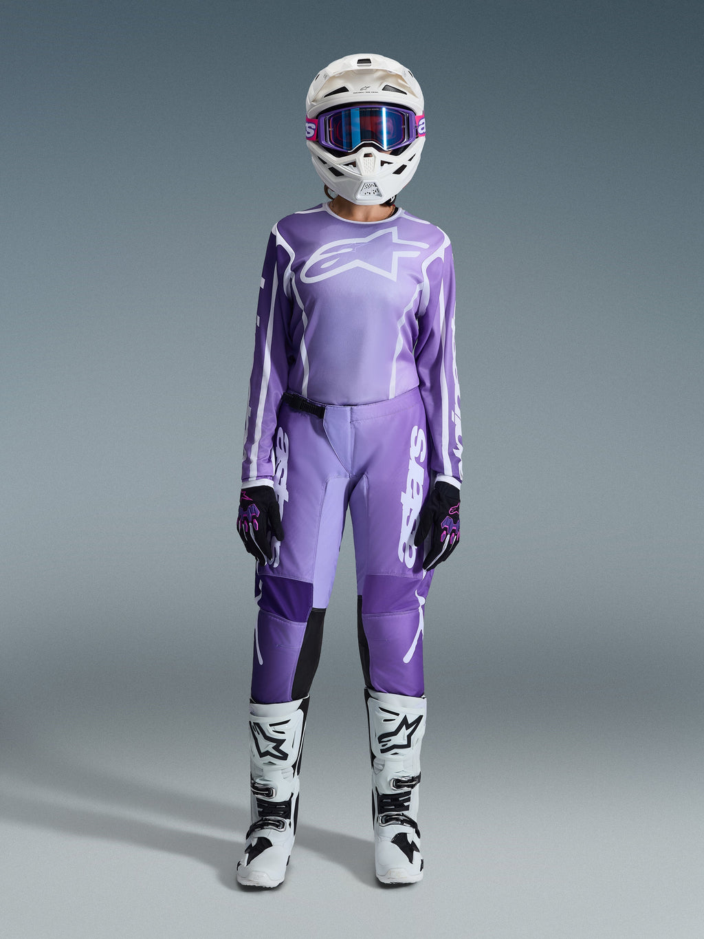 Pilota di motocross con completo Alpinestars Stella Fluid Apex 2026, maglia viola e bianca con pantaloni coordinati, casco bianco con occhiali rosa e stivali da fuoristrada bianchi e neri, kit da gara completo per motocross femminile.